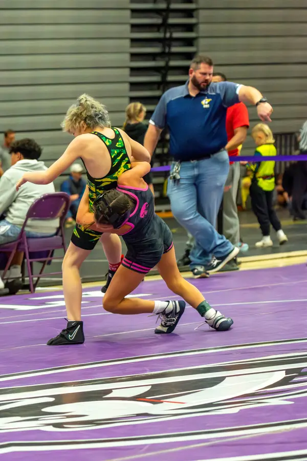 A wrestler in pink-trimmed gear drives into an opponent as the referee watches