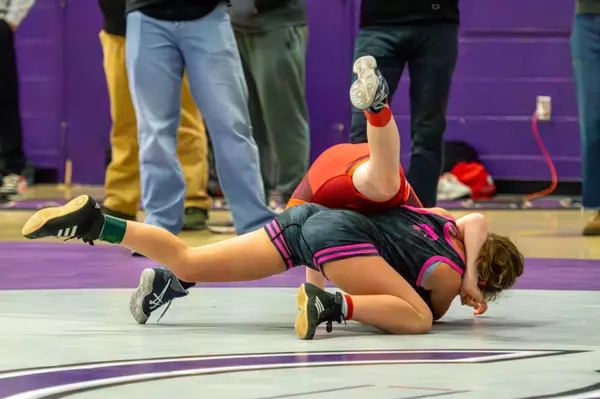 A wrestler in pink drives through a takedown, sending an opponent's legs into the air
