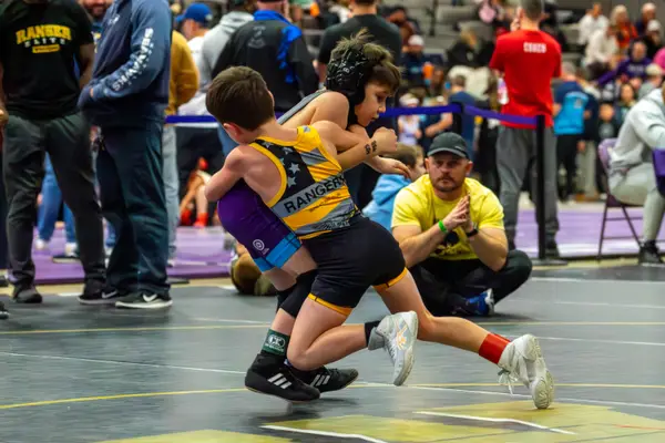 A young Rangers wrestler drives an opponent across the mat as a coach watches