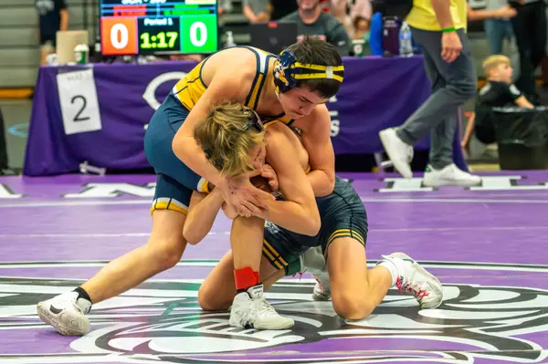 Two wrestlers lock up on a purple mat near the scorer's table