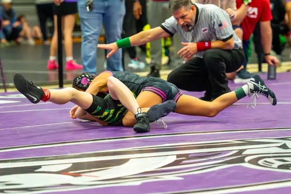A wrestler in pink headgear controls an opponent as the referee signals