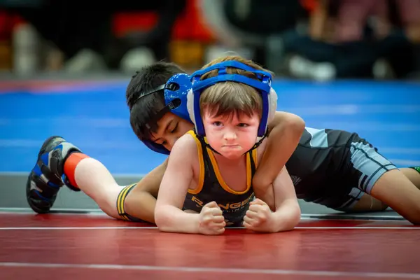A young Rangers wrestler grimaces on a red mat while an opponent holds on from behind