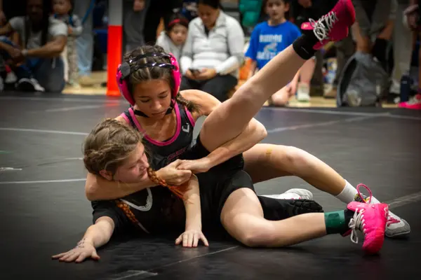 A Rangers wrestler in pink headgear rides an opponent on a black mat