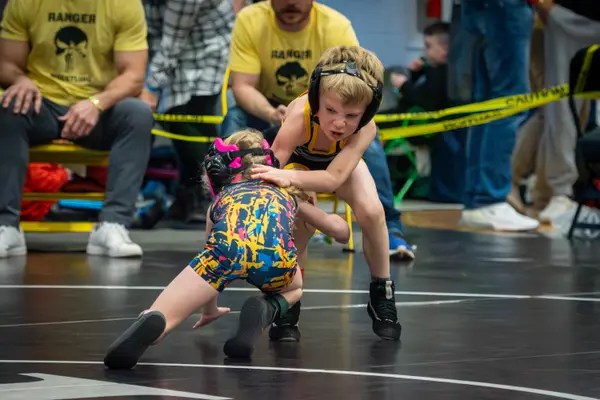 A young Rangers wrestler ties up with an opponent as coaches in Rangers shirts watch