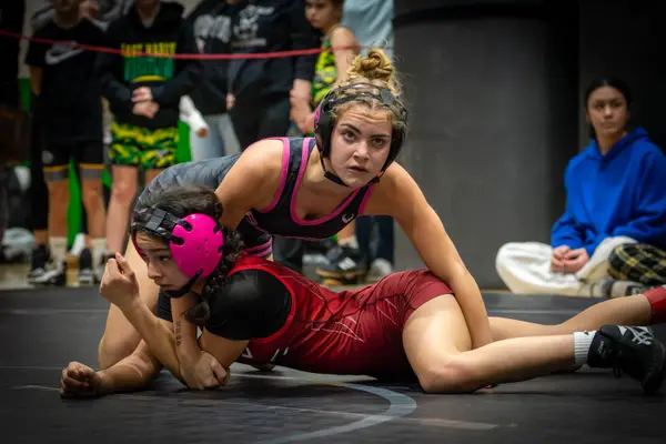 A wrestler in pink headgear rides an opponent while looking toward the mat edge