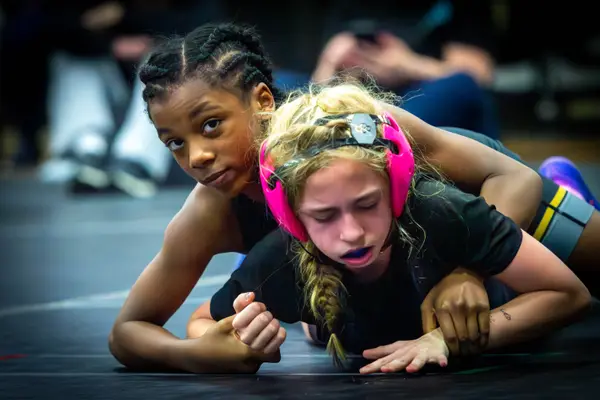 Two wrestlers on the mat, one in pink headgear looking down