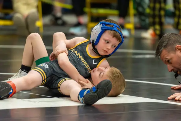 A young Rangers wrestler in blue headgear holds an opponent down as the referee checks