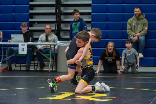 A young Rangers wrestler drives into an opponent as a parent cheers from the mat edge