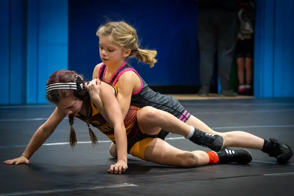 A young wrestler in pink-trimmed gear rides an opponent on a black mat