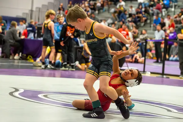 A Rangers wrestler steps over a falling opponent in a red singlet at a busy tournament